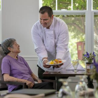 A chef serves a meal to a senior woman in a care home setting.