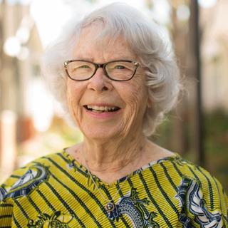 An elderly woman with glasses smiles warmly while standing outdoors.