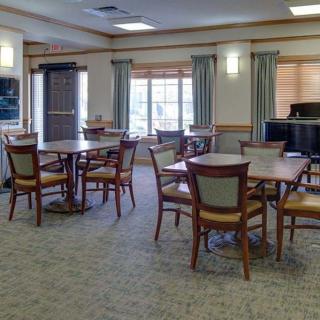 A well-lit dining area with multiple tables and chairs, featuring large windows with curtains and a carpeted floor.