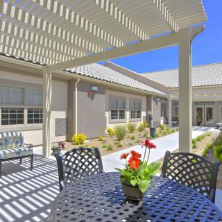 A sunny outdoor patio with a pergola providing shade over a table and chairs, adorned with red flowers.