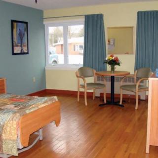 A well-lit hospital room with a bed, chairs, and a table, featuring wooden flooring and curtains.