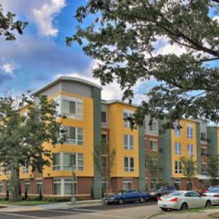 A modern apartment building surrounded by trees with parked cars along the street.