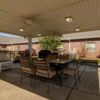 A cozy outdoor patio with a dining table and chairs under a covered area, adjacent to a brick building and garden.