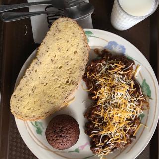 A meal featuring chili topped with cheese, a slice of bread, and a muffin alongside a cup of milk.