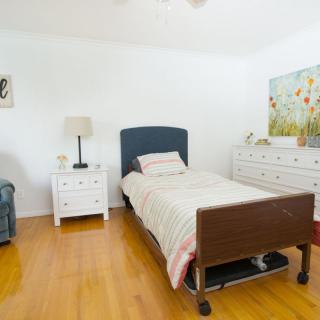 A bright bedroom with a single bed, nightstand, lamp, and dresser on a wooden floor.