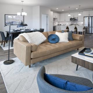A modern open-concept living space featuring a beige sofa with blue accent pillows, adjoining a kitchen and dining area.
