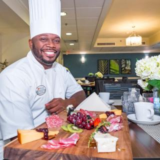 A smiling chef presents a beautifully arranged charcuterie board with various cheeses and fruits in a restaurant setting.