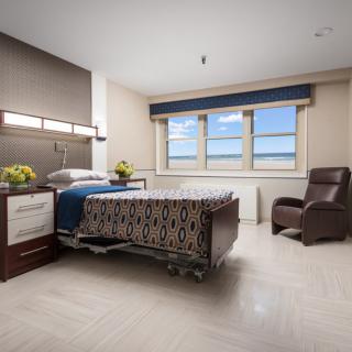 A clean and bright hospital room featuring a bed, a chair, and a large window.