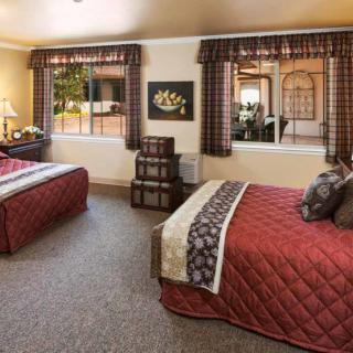 A cozy bedroom featuring two beds with red quilted bedding, large windows with patterned curtains, and a carpeted floor.