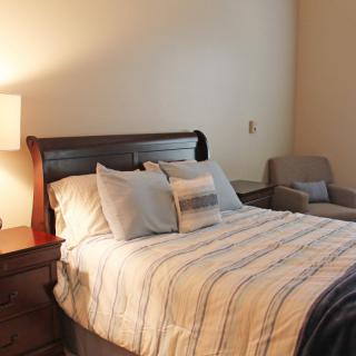 A cozy bedroom with a neatly made bed, an armchair, and a bedside lamp.