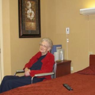 An elderly woman in a red jacket sitting in a wheelchair inside a room with artwork on the wall.