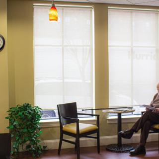 An elderly person sitting in a quiet waiting room with large windows and simple furnishings.