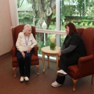An elderly woman and a caregiver sitting in chairs, engaging in conversation near a window.