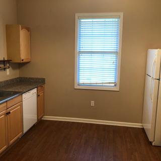 An empty kitchen with wooden cabinets, a refrigerator, dishwasher, and a window with blinds.