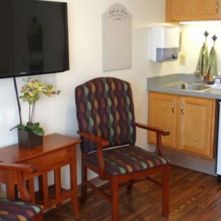 A cozy living space with a patterned armchair, wooden table, wall-mounted television, and a small kitchenette.