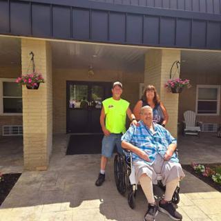 A joyful family moment captured on the porch with a grandparent in a wheelchair, flanked by two young family members.