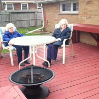 Two elderly individuals sitting on a wooden deck with patio furniture and a fire pit.