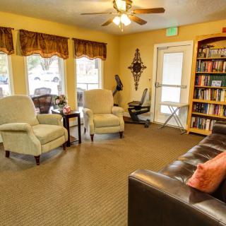 A cozy living room with comfortable seating and a bookshelf, featuring large windows and a ceiling fan.