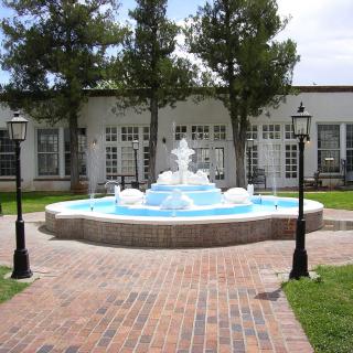 A tranquil courtyard with a central fountain, surrounded by lampposts and trees, adjacent to a building.