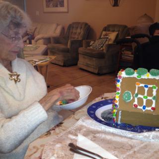 An elderly woman is sitting at a table beside a decorated gingerbread house, engaging in some craftwork indoors.