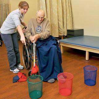 An elderly person in a wheelchair is assisted by a caregiver during a physical therapy session involving colorful baskets and bean bags.