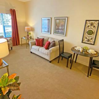 A cozy living room with a sofa, decorative pillows, and artworks on the wall. There's a small dining table with two chairs and a window with red curtains providing natural light.