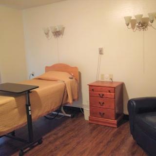 A simple hospital room with a bed, wooden nightstand, and comfortable seating.