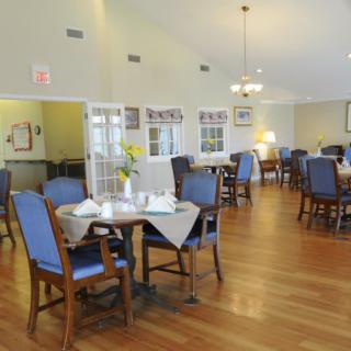 A cozy dining room with neatly arranged tables and blue cushioned chairs, featuring warm lighting and wooden floors.