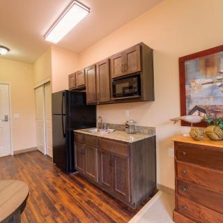 A compact kitchen with dark wood cabinets, a black refrigerator and microwave, and wooden flooring.