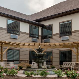 A peaceful courtyard with a fountain, pergola, and benches surrounded by a two-story building.