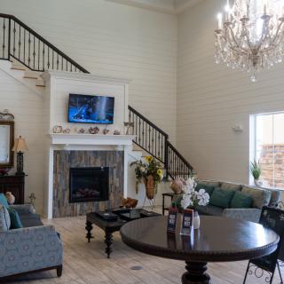 A spacious living room with high ceilings, featuring a grand chandelier, a television mounted above the fireplace, and comfortable seating around a coffee table.
