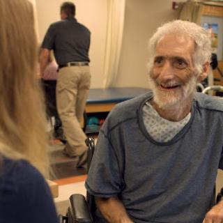 An elderly man smiling during a conversation with a caregiver in a rehabilitation setting.