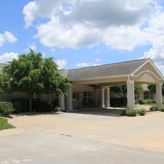 A view of a building entrance with a driveway, surrounded by trees under a partly cloudy sky.
