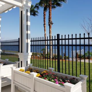 A scenic garden with colorful flowers overlooks a tranquil sea, framed by palm trees and a black metal fence.