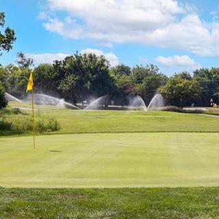 A well-maintained golf course featuring a green with sprinklers in operation under a clear blue sky.