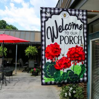A cheerful 'Welcome to Our Porch' sign adorned with vibrant red flowers, inviting guests to enjoy the outdoor seating area.