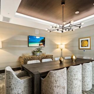 A modern dining room featuring a large table with upholstered chairs, a stylish chandelier, and decorative elements including wall art and a television.