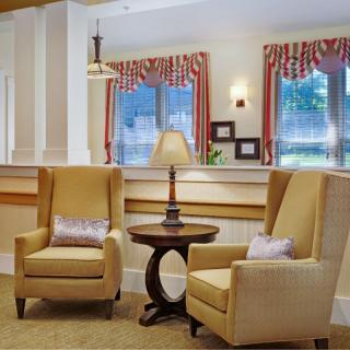 A cozy lounge area with two armchairs and a table topped with a lamp, framed by windows with red and white curtains.