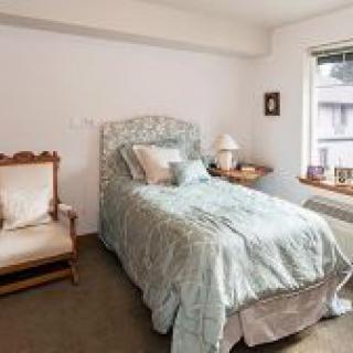 A cozy bedroom with a single bed, adorned with decorative pillows, an armchair, and a bedside lamp by the window.