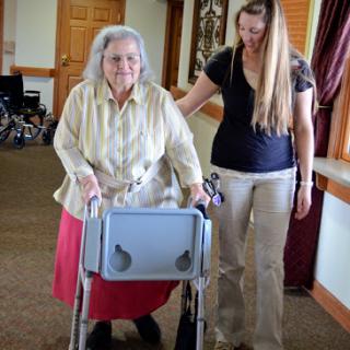 An elderly woman is assisted by a caregiver using a walker in a community setting.