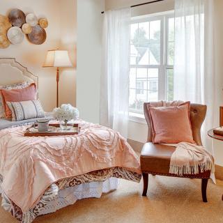 A cozy bedroom with soft colors and elegant decor, featuring a bed, a comfortable chair, and large windows with sheer curtains.