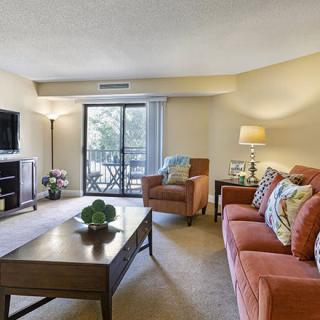 A cozy living room with a sofa, armchair, and coffee table, featuring a TV and sliding glass doors leading to a balcony.