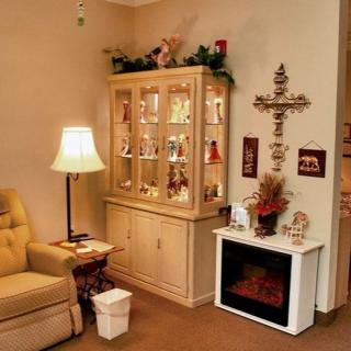 A cozy living room corner with a display cabinet, comfortable chair, and decorative cross above an electric fireplace.