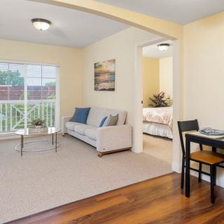 A cozy living room with a sofa, coffee table, and a small dining area featuring wood flooring and a large window.