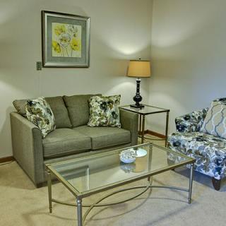 A cozy living room with a sofa, patterned chair, glass coffee table, and a lamp beside artwork.