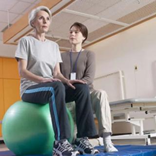 An elderly woman is guided by a trainer during a physical therapy session on an exercise ball.