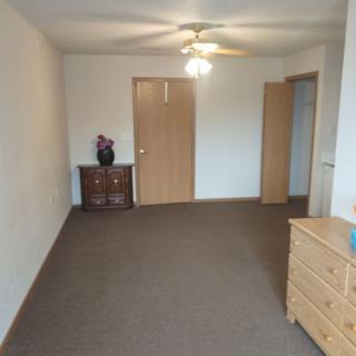 A simple living room with minimal furniture, including a ceiling fan, carpeted floor, and wooden doors.