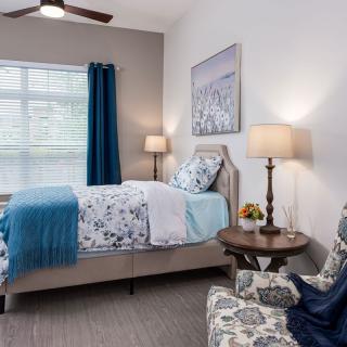 A cozy bedroom with a blue and white theme, featuring a bed, bedside lamp, armchair, and window with curtains.
