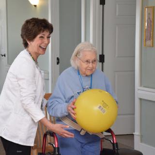 A caregiver joyfully assists an elderly woman as she engages in a fitness activity using a yellow ball.