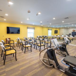 A modern exercise room features rows of chairs, fitness equipment, and a television mounted on a yellow wall.